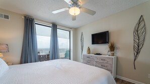 A bedroom with a large bed, a ceiling fan, a flat-screen TV, and a view of the outdoors. - Pelican Pointe 504 by Liquid Life (Orange Beach)