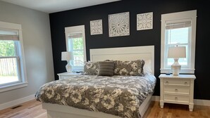 A bedroom with a large bed, a nightstand, a dresser, and two windows with blinds.