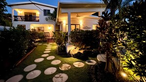 Een huis met twee verdiepingen en een goed verlichte tuin met een pad van stenen en diverse planten.