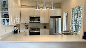 Een moderne keuken met witte kasten, roestvrijstalen apparatuur en hangende lampen. Er staan drie houten krukken en een kleurrijk geweven vloerkleed.
