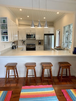 A modern kitchen with white cabinets, stainless steel appliances, and pendant lighting. There are three wooden stools and a colorful woven rug on the floor.