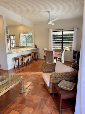 A living room with a wicker sofa, a glass coffee table, and a TV. There is a kitchen area with a counter and stools.