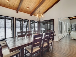 Un comedor con mesa y sillas de madera, ventanas amplias con vista a árboles y un paisaje nevado, y una araña.