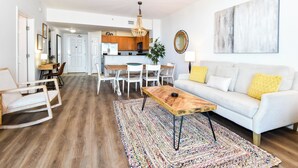 A modern living room with a white sofa, a wooden coffee table, and a colorful rug. There is a dining area with white chairs and a table, and a kitchen in the background. - Fully Updated! Beautiful 2 Bedroom Condo with Seasonal Beach Shuttle! (Destin)