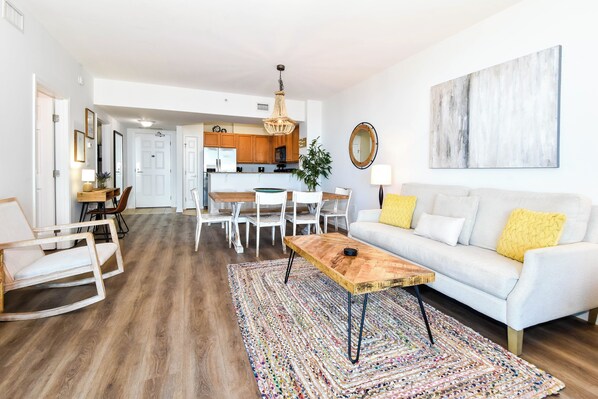 Una sala de estar moderna con un sofá blanco, una mesa de centro de madera y un tapiz colorido. Se observa un comedor con sillas blancas y una mesa, y una cocina al fondo.