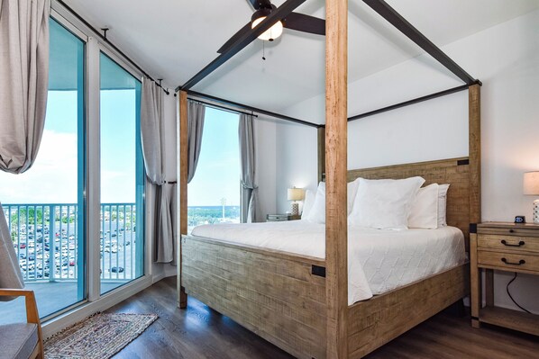 Uma cama de dossel com cabeceira de madeira, uma mesa de cabeceira com abajur, e vista para um parque de estacionamento através de janelas amplas.