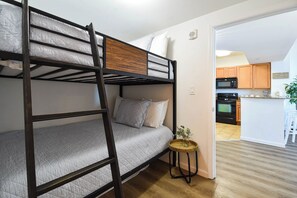 Uma cama beliche com cabeceira de madeira, uma pequena mesa redonda com uma planta e uma cozinha ao fundo.