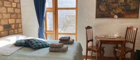 A room with a bed, a table, and chairs. There is a window with curtains and a painting on the wall.