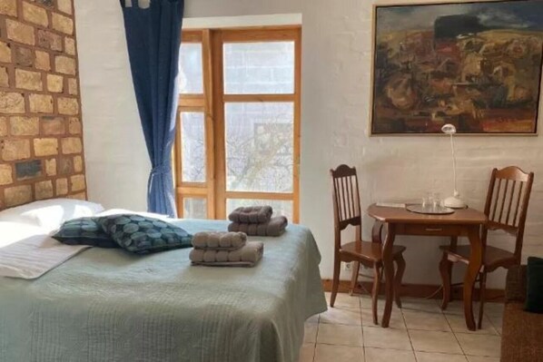 Une pièce comprenant un lit, une table et des chaises. Il y a une fenêtre avec des rideaux et un tableau accroché au mur.