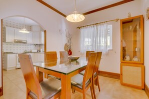 A dining area with a glass-top table and wooden chairs, a kitchen in the background, and a decorative cabinet with glass doors.