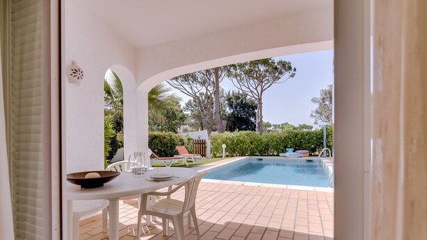 A patio with a pool, white furniture, and a view of trees and greenery.