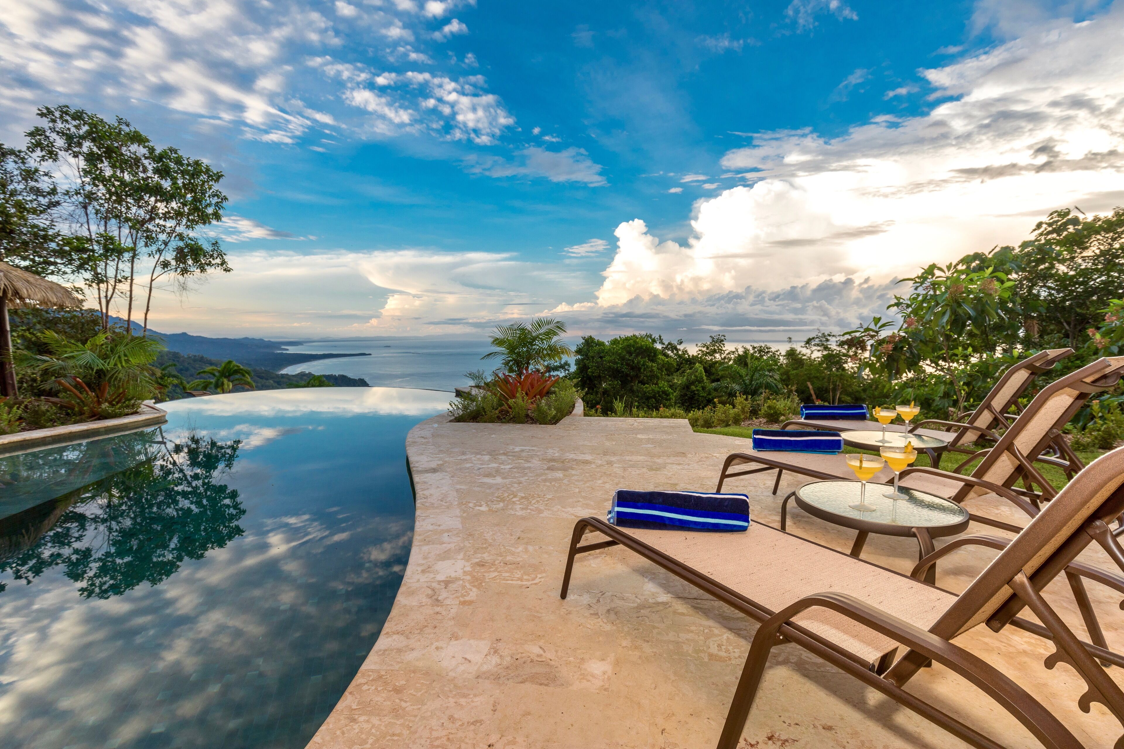 Área à beira da piscina com espreguiçadeiras e uma mesa, com vista para um corpo d'água e montanhas.