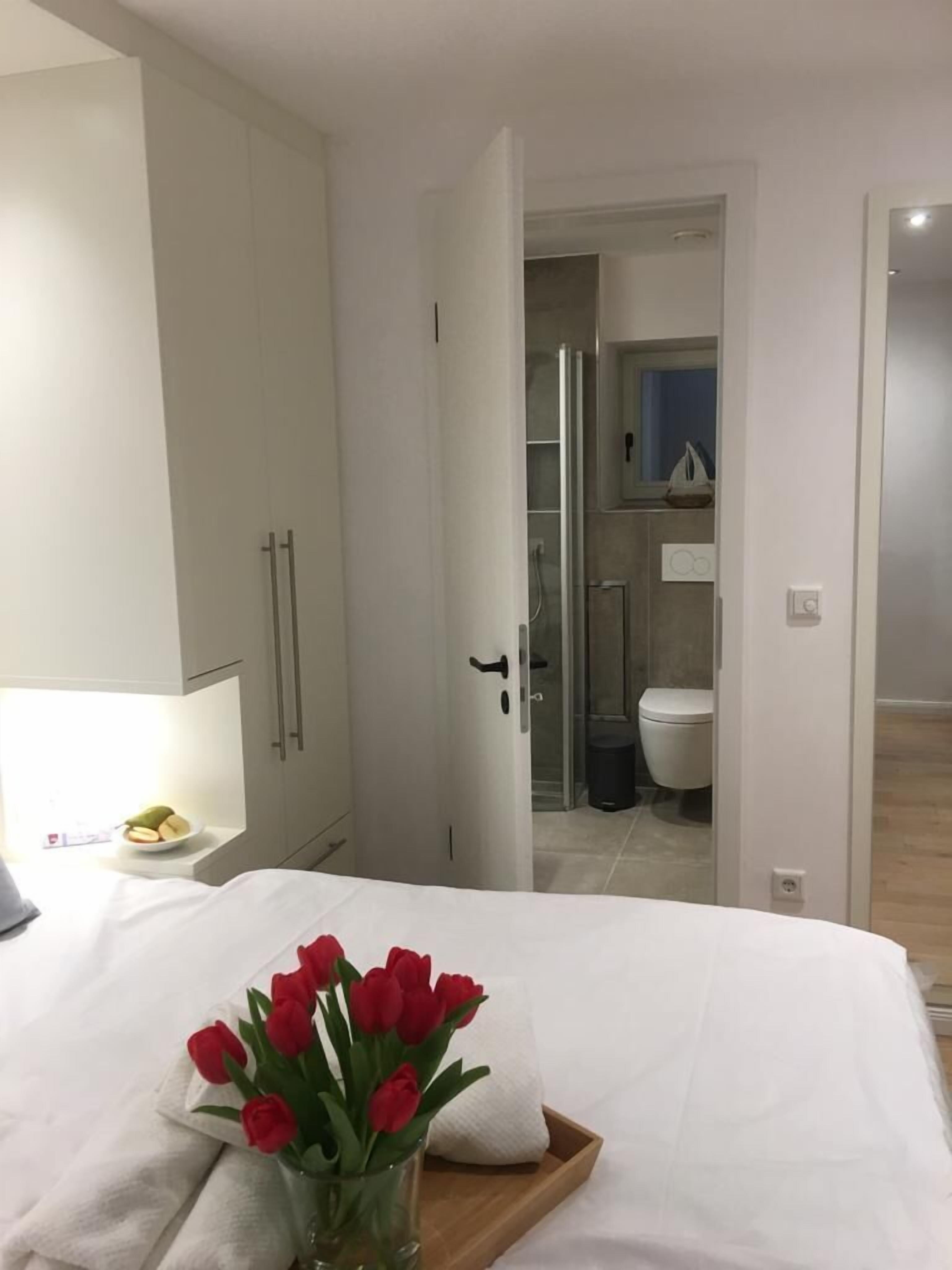 A bedroom with a bed, a nightstand, a vase with red flowers, and a view into the bathroom.