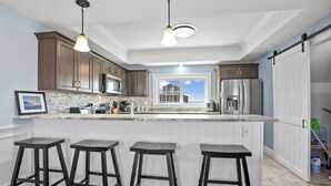 A modern kitchen with a large island, bar stools, stainless steel appliances, and a view to the outside. - At the Water's Edge by Seaside Vacations & Sales (Chincoteague Island)