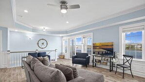 A spacious living room with a grey sofa, a blue armchair, and a dining area with a TV displaying a sunset view. - At the Water's Edge by Seaside Vacations & Sales (Chincoteague Island)