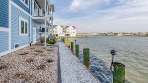 A waterfront property with a gravel path leading to a dock, a blue building with a balcony, and a white building in the background. - At the Water's Edge by Seaside Vacations & Sales (Chincoteague Island)