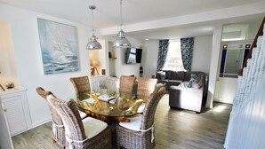 A dining area with wicker chairs and a glass-top table, a living room with a sofa and a television, and a staircase with white railings. - Fantastic location in the harbour bowl (Brixham)