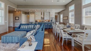 A spacious living area with a large blue sectional sofa, a dining table with white chairs, and a kitchen in the background.