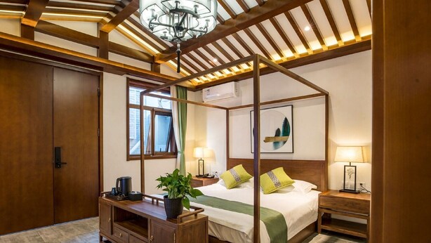 A four-poster bed with a green and white bedspread, a wooden nightstand with a lamp, and a potted plant on a wooden console table.