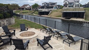 A patio area with outdoor seating and a fire pit, overlooking a water feature and modern houses.