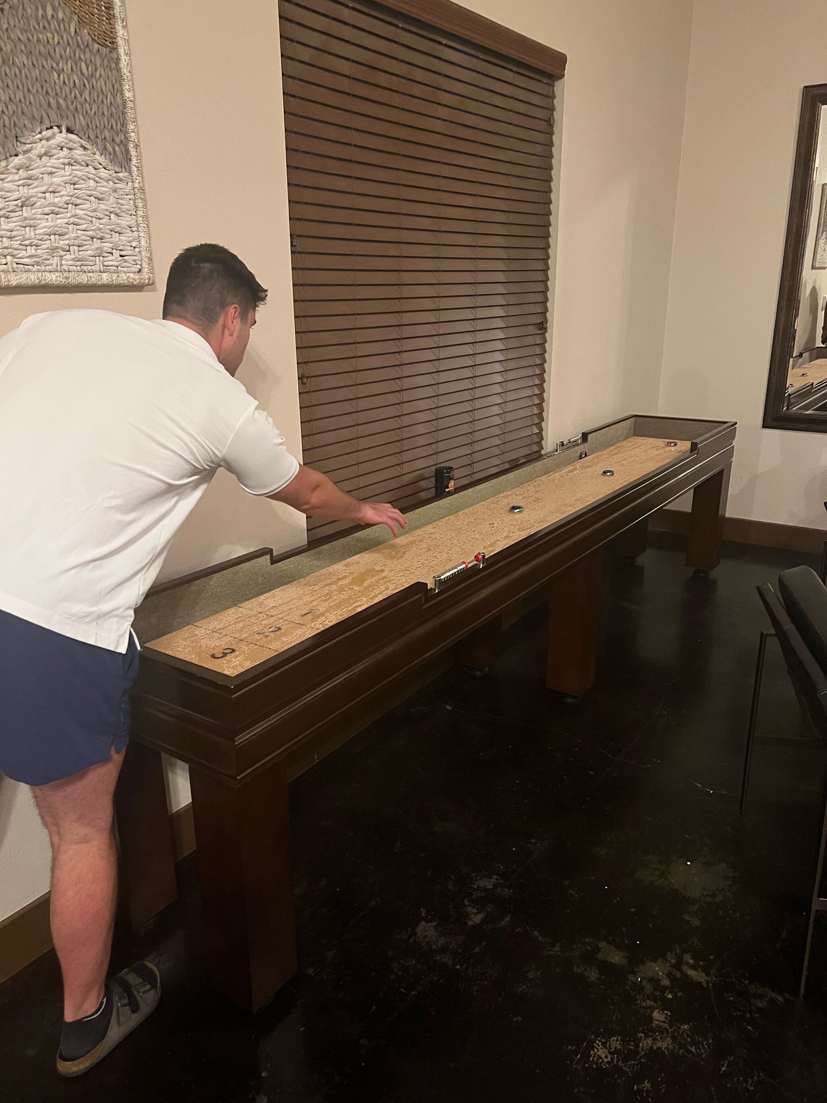 A pool table in a room with a dark floor, a window with blinds, and a framed picture on the wall.