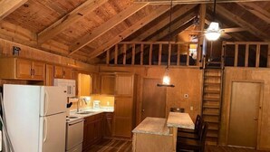 Interno rustico di una baita con pareti e soffitto in legno, un angolo cottura con frigorifero, fornello e microonde, una zona pranzo con tavolo e sedie, e una scala che conduce a un piano superiore.