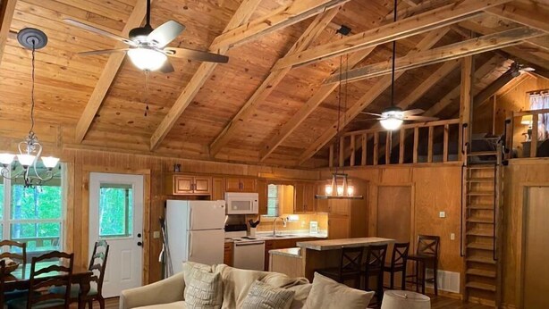 Interno accogliente di una baita con soffitto in legno, zona pranzo, cucina e soggiorno con divano e tavolino.