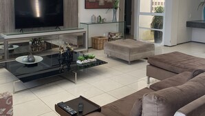 Ett modernt vardagsrum med en platt-TV, ett soffbord i glas, en brun soffa och ett stort fönster.
