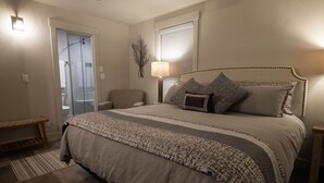 Une chambre à coucher avec un grand lit, une chaise, une table de chevet, une lampe et une porte donnant sur une salle de bain.