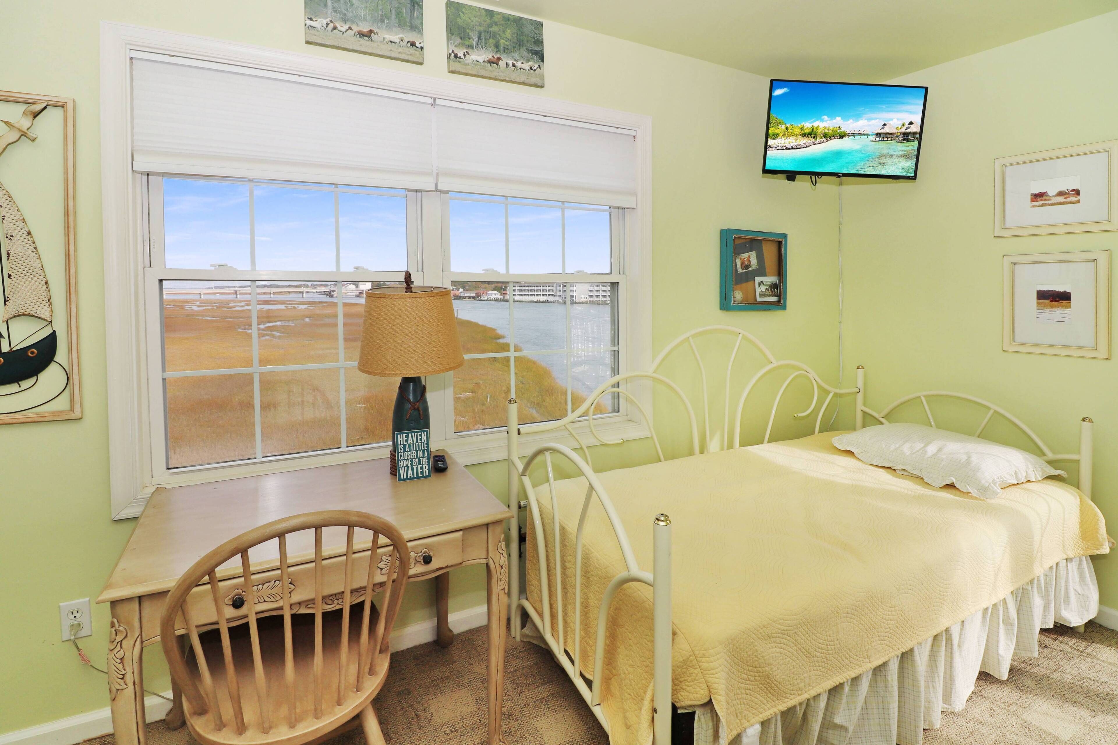 A bedroom with a bed, a desk, a chair, a window with a view of a beach, and a television mounted on the wall.