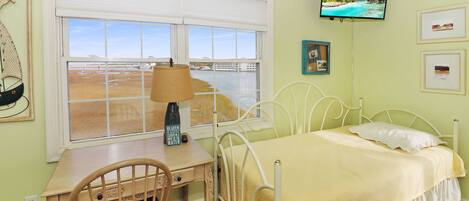 A bedroom with a bed, a desk, a chair, a window with a view of a beach, and a television mounted on the wall.