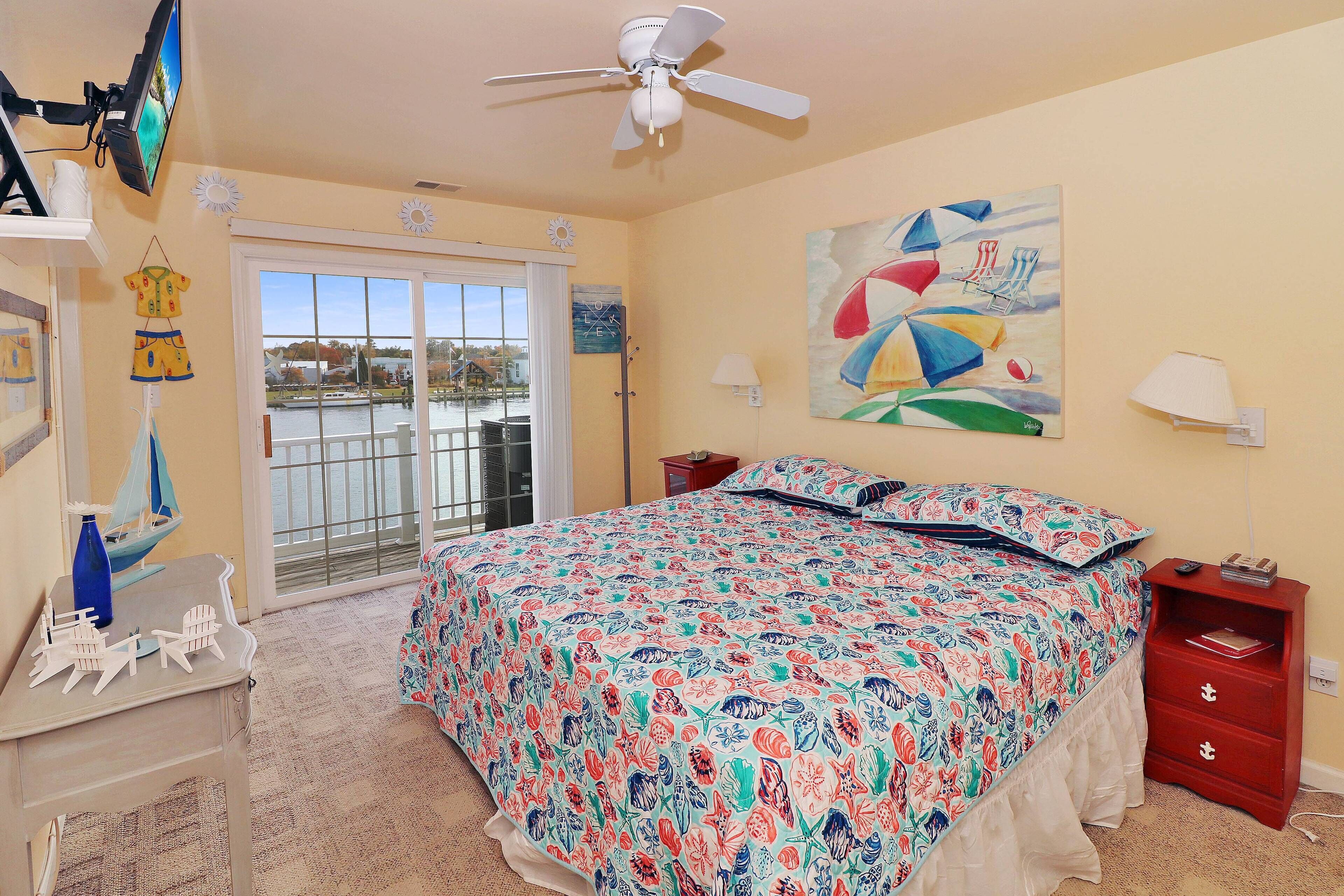 A bedroom with a large bed, a ceiling fan, a TV, and a view of the outdoors through a sliding door.