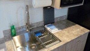 Una cocina con un lavamanos de acero inoxidable, un escurreplatos, un microondas y una botella de detergente.