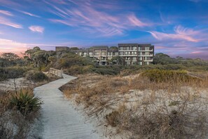 Un hotel frente a la playa con varios pisos, un paseo marítimo de madera que lleva a la entrada y vegetación natural circundante.