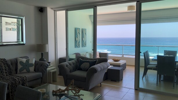 Un salón moderno con un ventanal de vidrio que da a un balcón con vistas al océano.