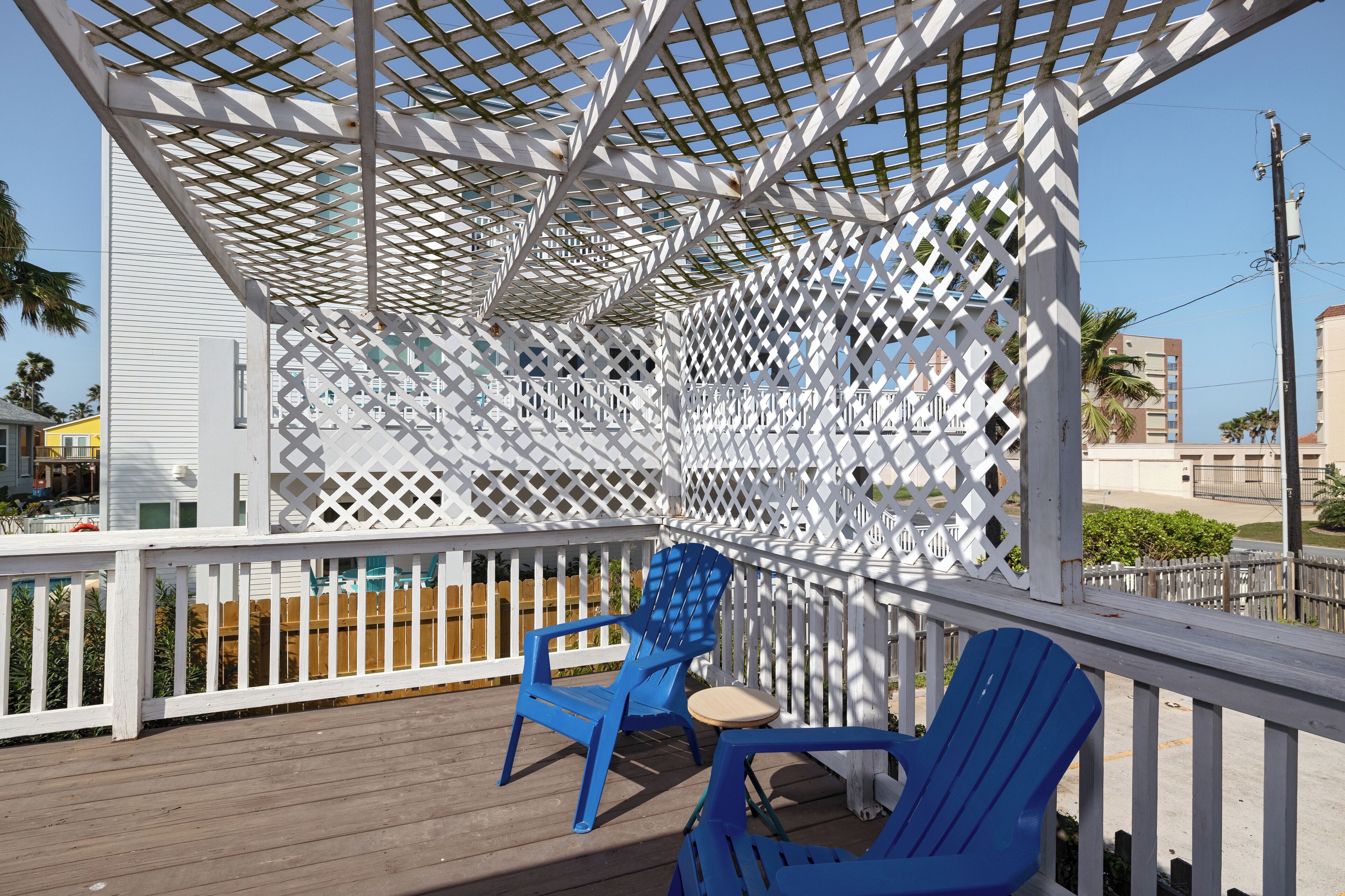 A covered outdoor deck with a lattice structure, blue Adirondack chairs, and a small table.