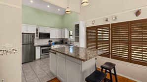 Una cocina con armarios blancos, encimera de granito, refrigerador de acero inoxidable y microondas. Hay dos lámparas colgantes, un taburete de madera y una ventana con persianas.