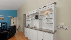 Cocina con armarios blancos, encimeras de granito y estantes de vidrio. Se observa una silla de cuero negro y una lámpara fijada a la pared.
