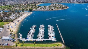 Vista aérea de un puerto deportivo con barcos amarrados, una zona de playa con sombrillas y un paisaje urbano al fondo.