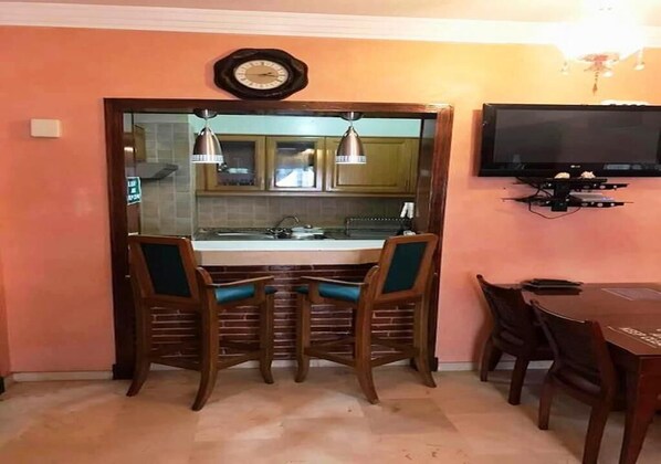 Une cuisine avec un comptoir en bois, deux chaises dotées de coussins turquoise, une horloge et un téléviseur fixé au mur.