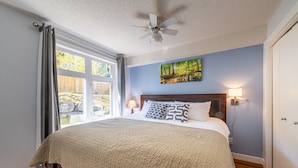 A bedroom with a large bed, a ceiling fan, a window with curtains, a painting on the wall, and a nightstand with a lamp. - 4 Bedrooms Suite - Group of Families (Canmore)