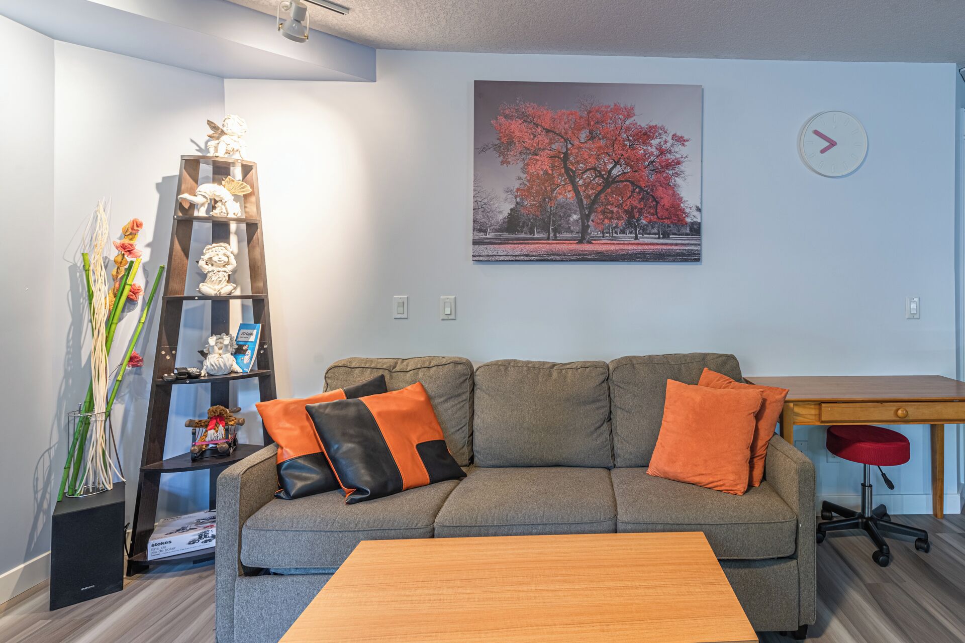 A living room with a grey sofa, orange and black cushions, a wooden coffee table, a bookshelf with decorative items, a painting of a tree, and a clock on the wall.