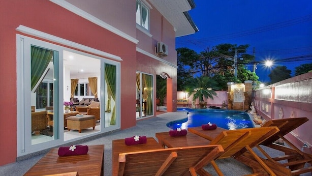 Zona de piscina con sillones de madera y una pileta, rodeada por un edificio rosa con puertas de vidrio que dan a una sala de estar.