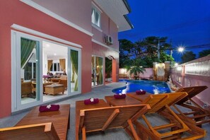 A poolside area with wooden lounge chairs and a pool, surrounded by a pink building with glass doors leading to a living room.