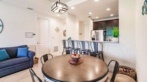 A dining area with a round table, chairs, and a bowl of fruit. There is a sofa with cushions in the background.
