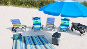 A beach setup with striped beach chairs, a blue umbrella, a stroller, and a cooler.
