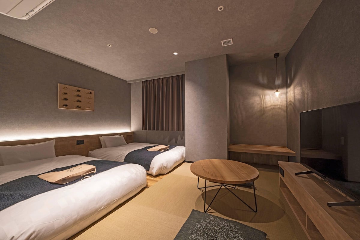 A modern hotel room with two beds, a wooden bench, a small round table, and a TV.