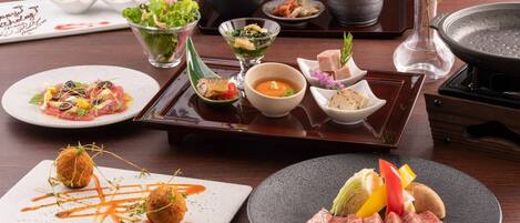 A variety of dishes on a wooden table, including sushi, sashimi, and a meat dish, with a teapot and a vase of flowers in the background.