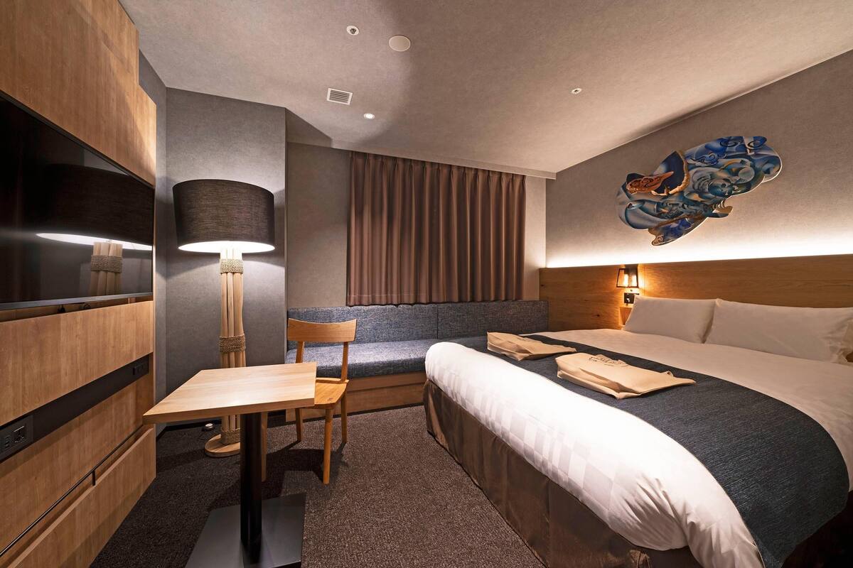 A hotel room with a bed, a desk, a chair, a lamp, and a wall-mounted artwork.