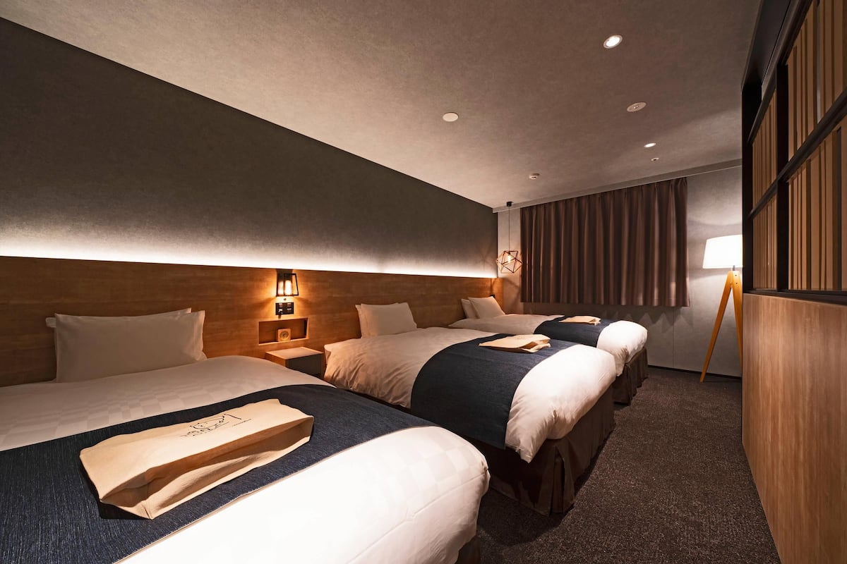 A hotel room with three beds, wooden headboard, and a standing lamp.